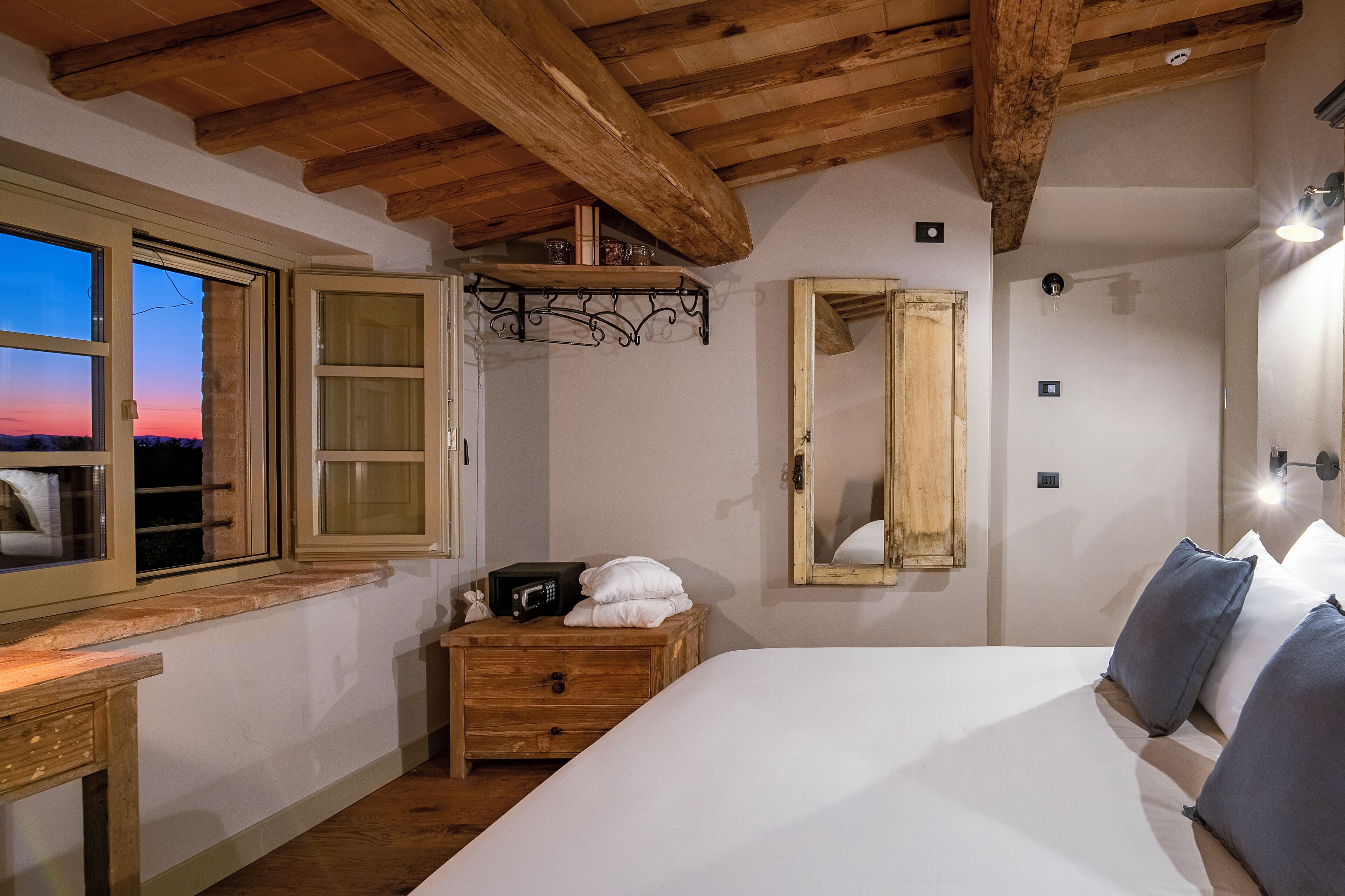 Une chambre avec un plafond en bois, un grand lit, une table de chevet avec une radio, une commode, un miroir et une fenêtre donnant sur le coucher de soleil.