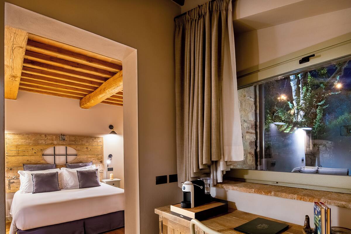 A bedroom with a wooden headboard, a bed with white and dark pillows, a bedside table with a coffee maker, and a window with curtains.