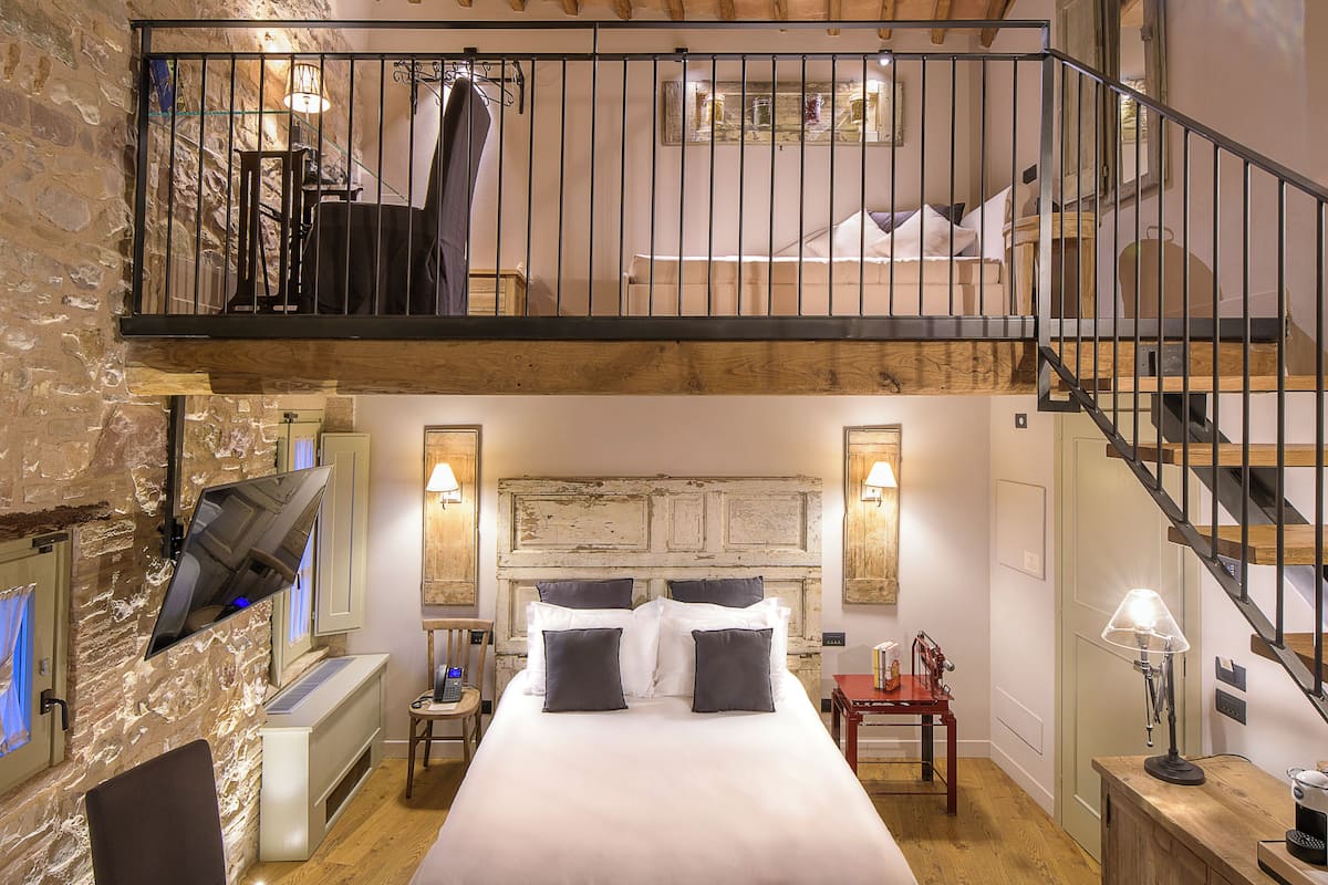 A bedroom with a large bed, a TV mounted on the wall, a wooden staircase, and a stone wall.