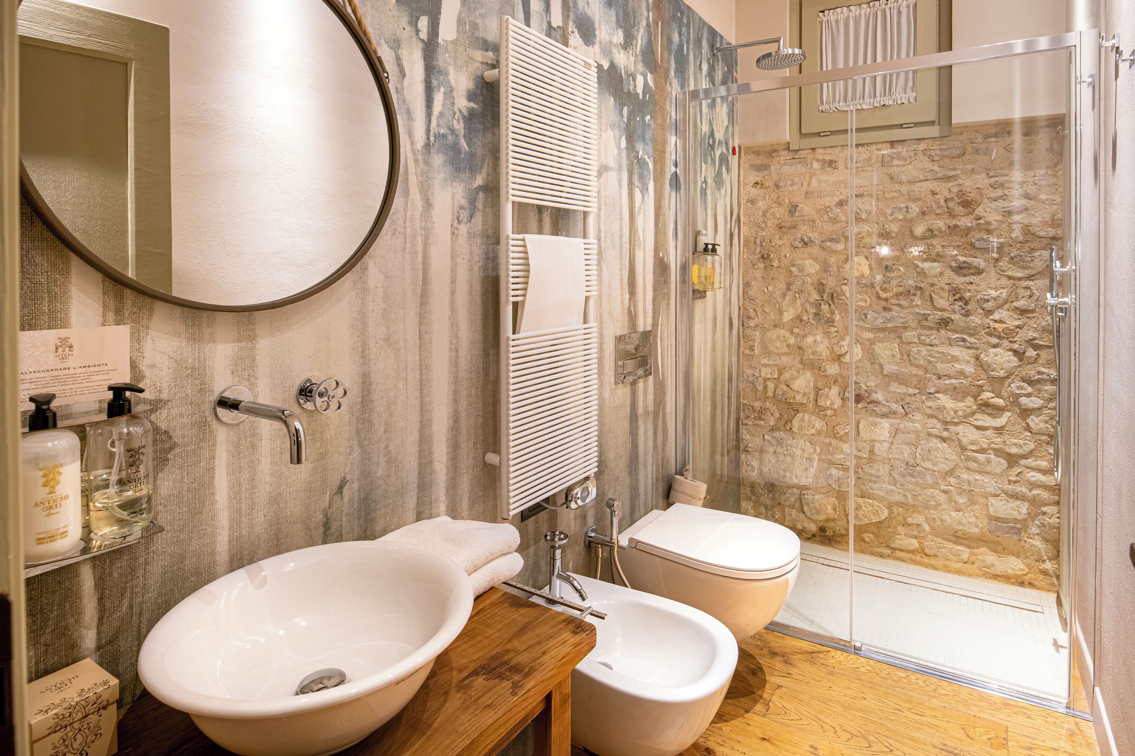 A bathroom with a wooden vanity, two sinks, a mirror, a towel rack, a shower with a stone wall, and a toilet.