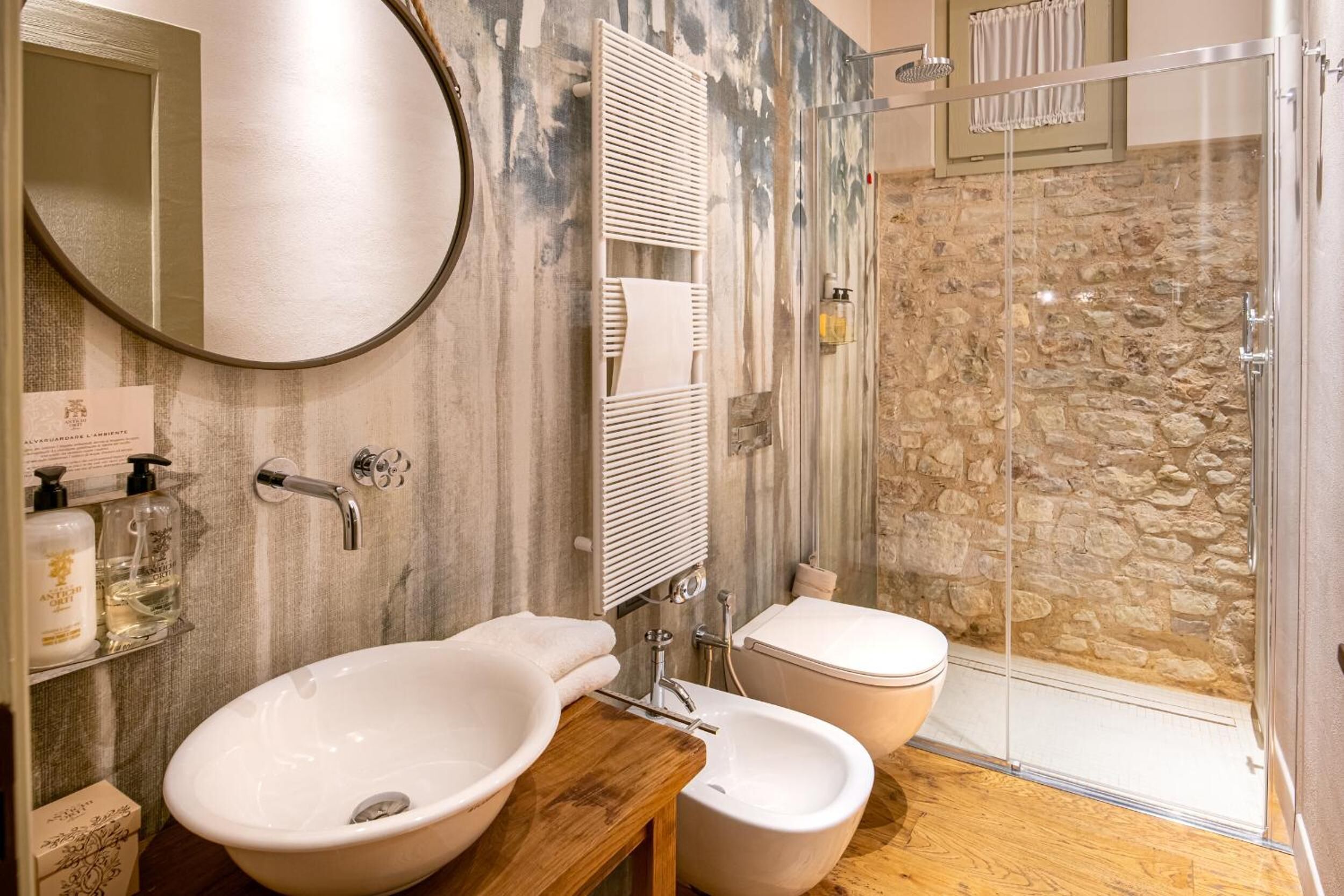 Ein Badezimmer mit rundem Spiegel, zwei Waschbecken, Toilette und Dusche mit Steinwand.
