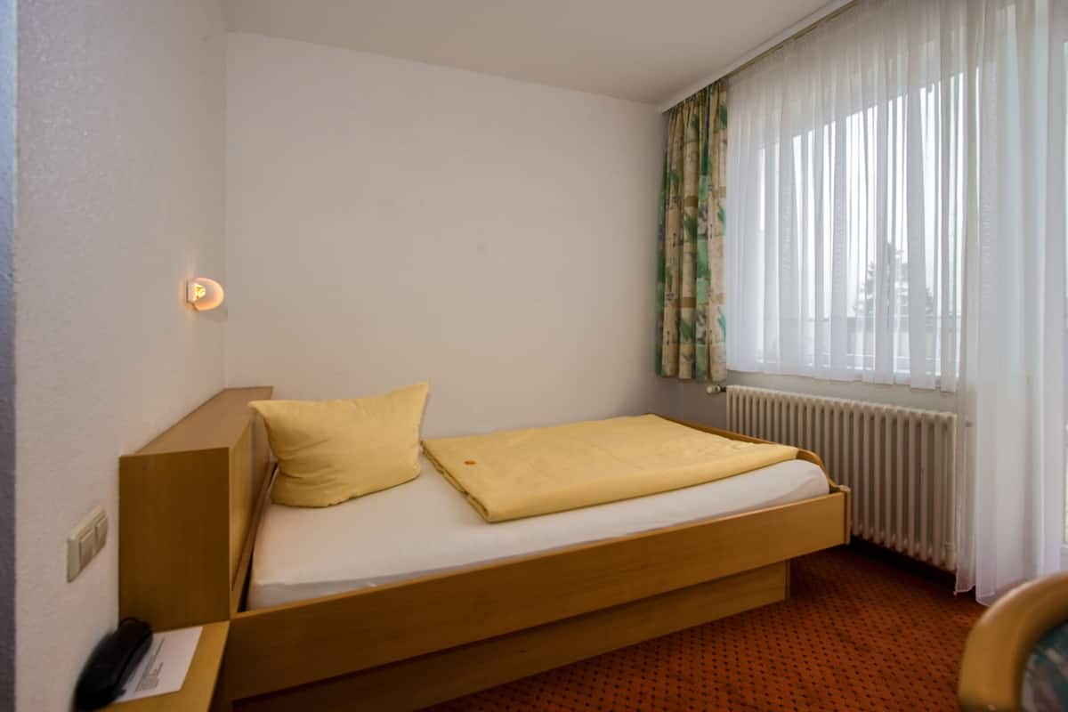Un lit simple avec un cadre en bois, un oreiller jaune et une couverture. Un radiateur se trouve sous une fenêtre ornée de rideaux légers.