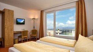 A hotel room with two beds, a desk, a chair, a TV, and a balcony with a view.