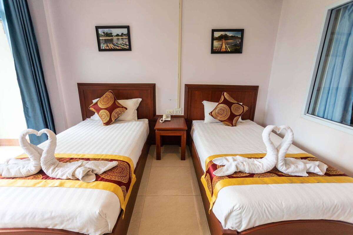 A hotel room with two beds, each with a heart-shaped towel arrangement, a small bedside table, and a wall-mounted phone.