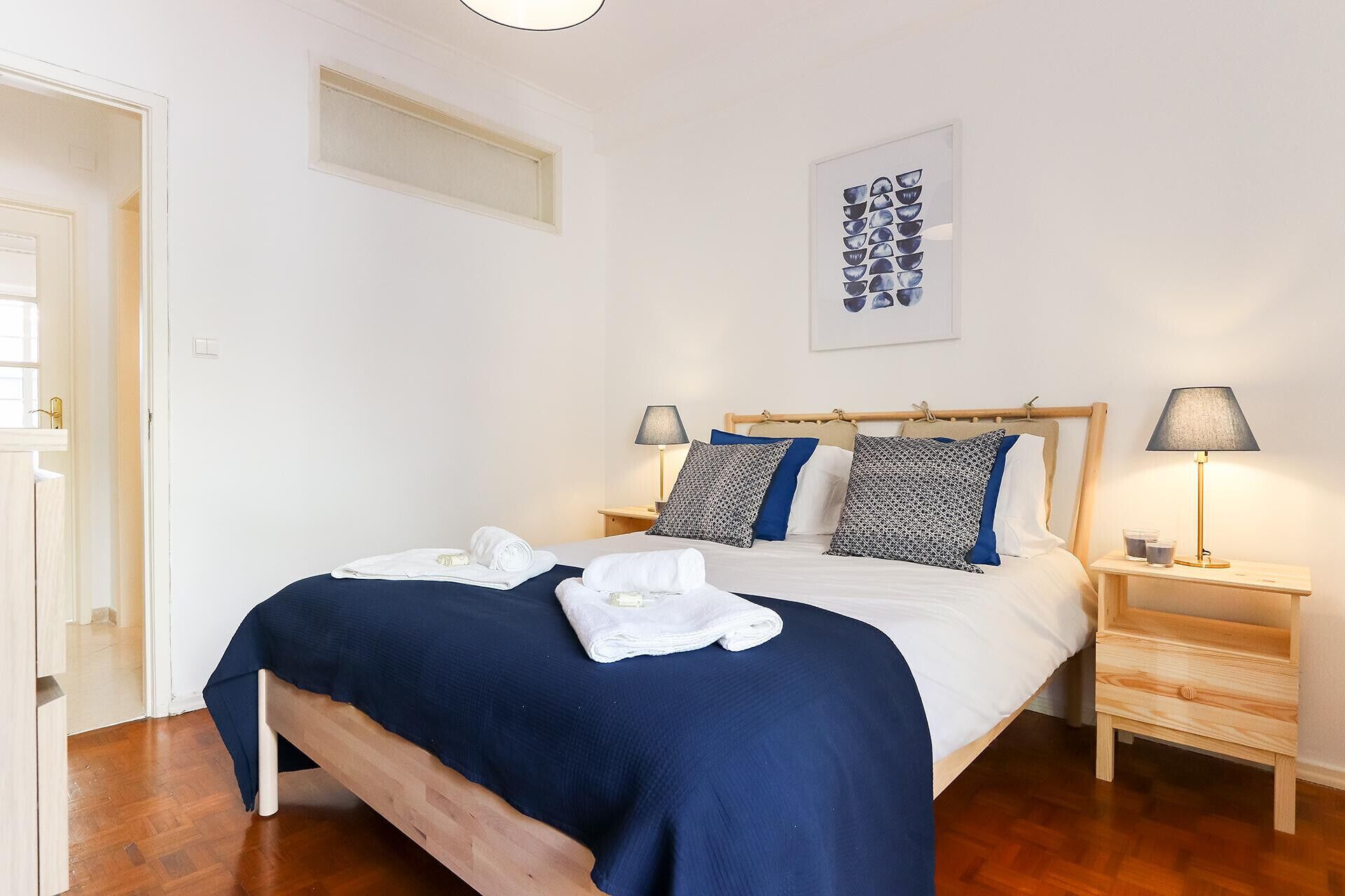 Een net opgemaakt bed met een blauwe sprei en witte lakens, twee bedlampen, een houten nachtkastje en een lijst met een kunstwerk aan de muur.