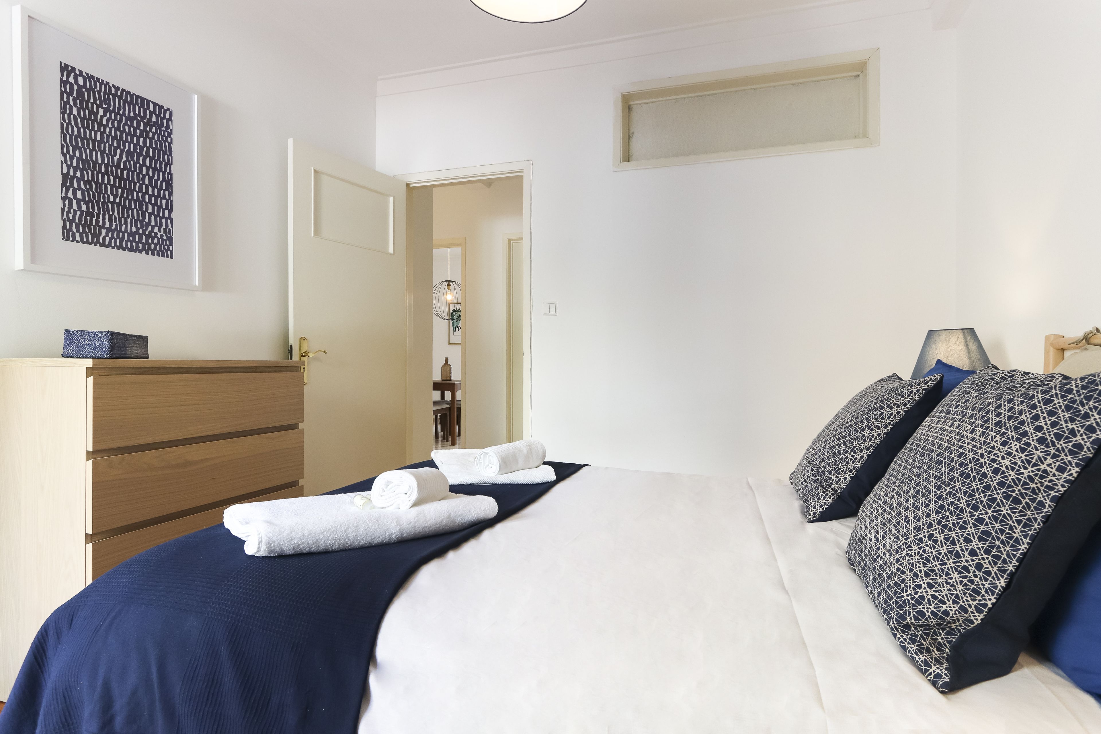Una cama bien hecha con ropa de cama blanca y azul marino, un aparador de madera y una obra de arte abstracta enmarcada en la pared.