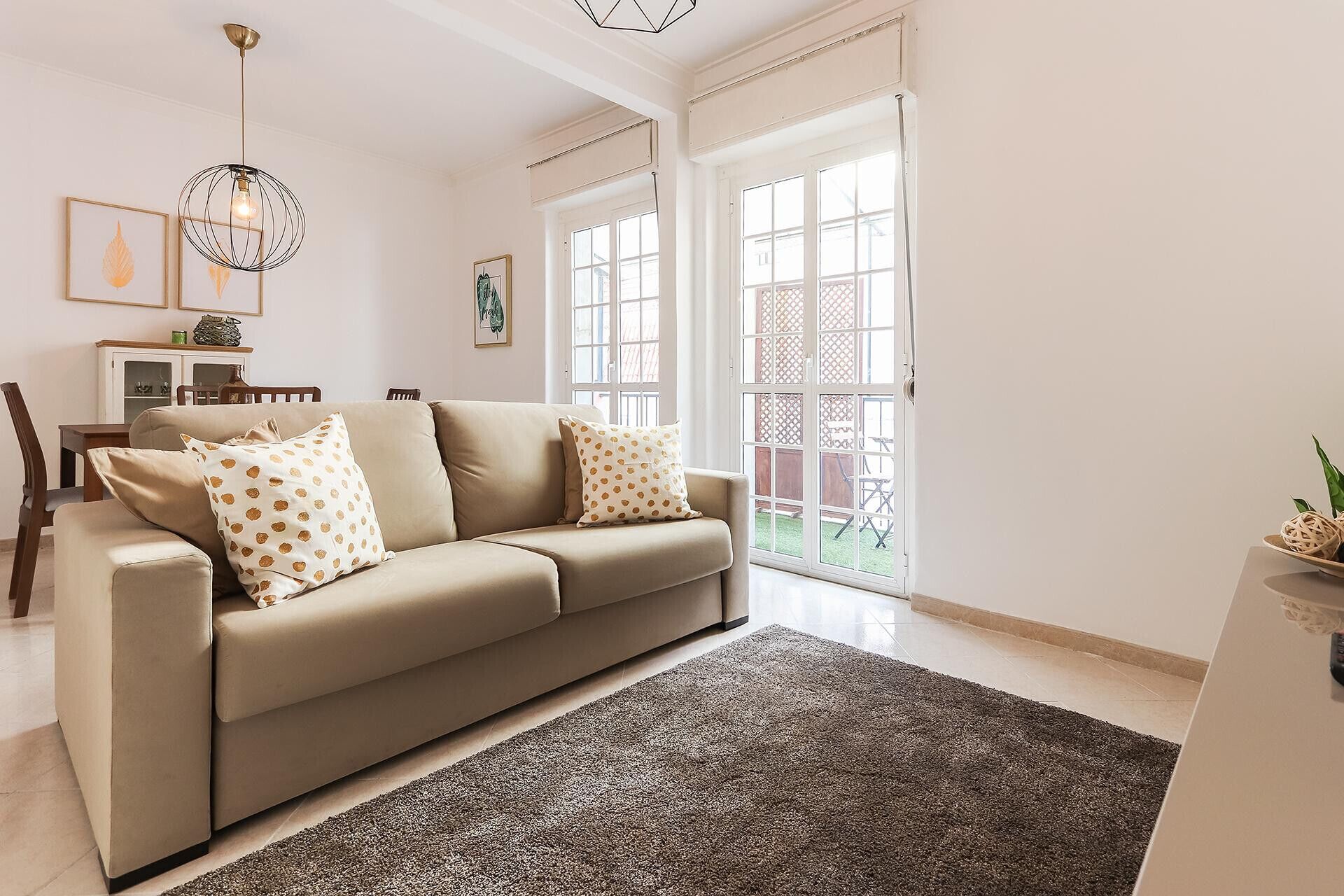 Ein Wohnzimmer mit einem beigefarbenen Sofa, gemusterten Kissen, einem dunklen Teppich und einem Essbereich mit Tisch und Stühlen.