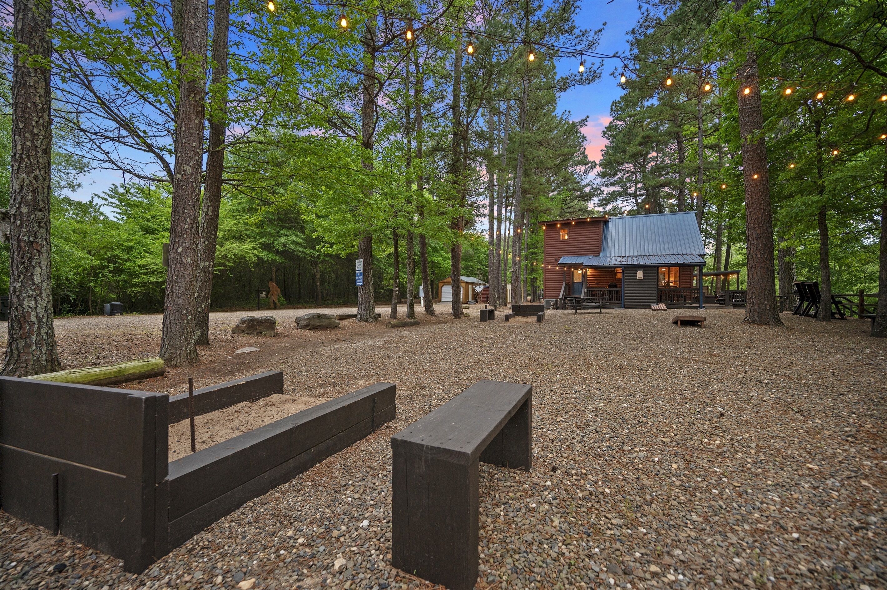 A rustic cabin with a gravel ground, wooden benches, and string lights.