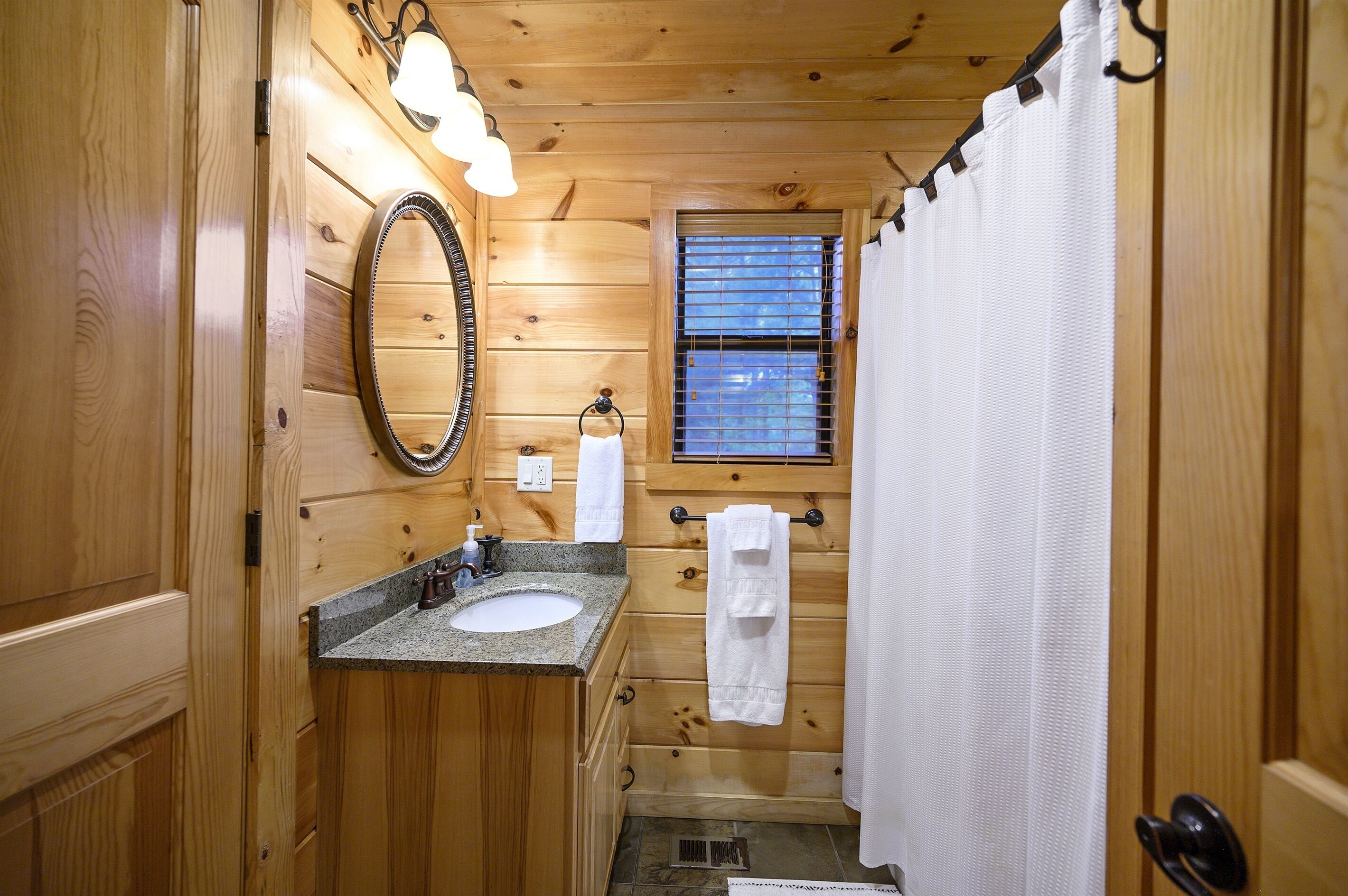 A wooden cabin bathroom with a granite sink, a mirror, a window with blinds, and a shower curtain.