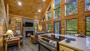 Ein gemütliches Blockhaus-Interieur mit Kamin aus Stein, Holzwänden, einer Couch, einem Fernseher und einer Küchenzeile.