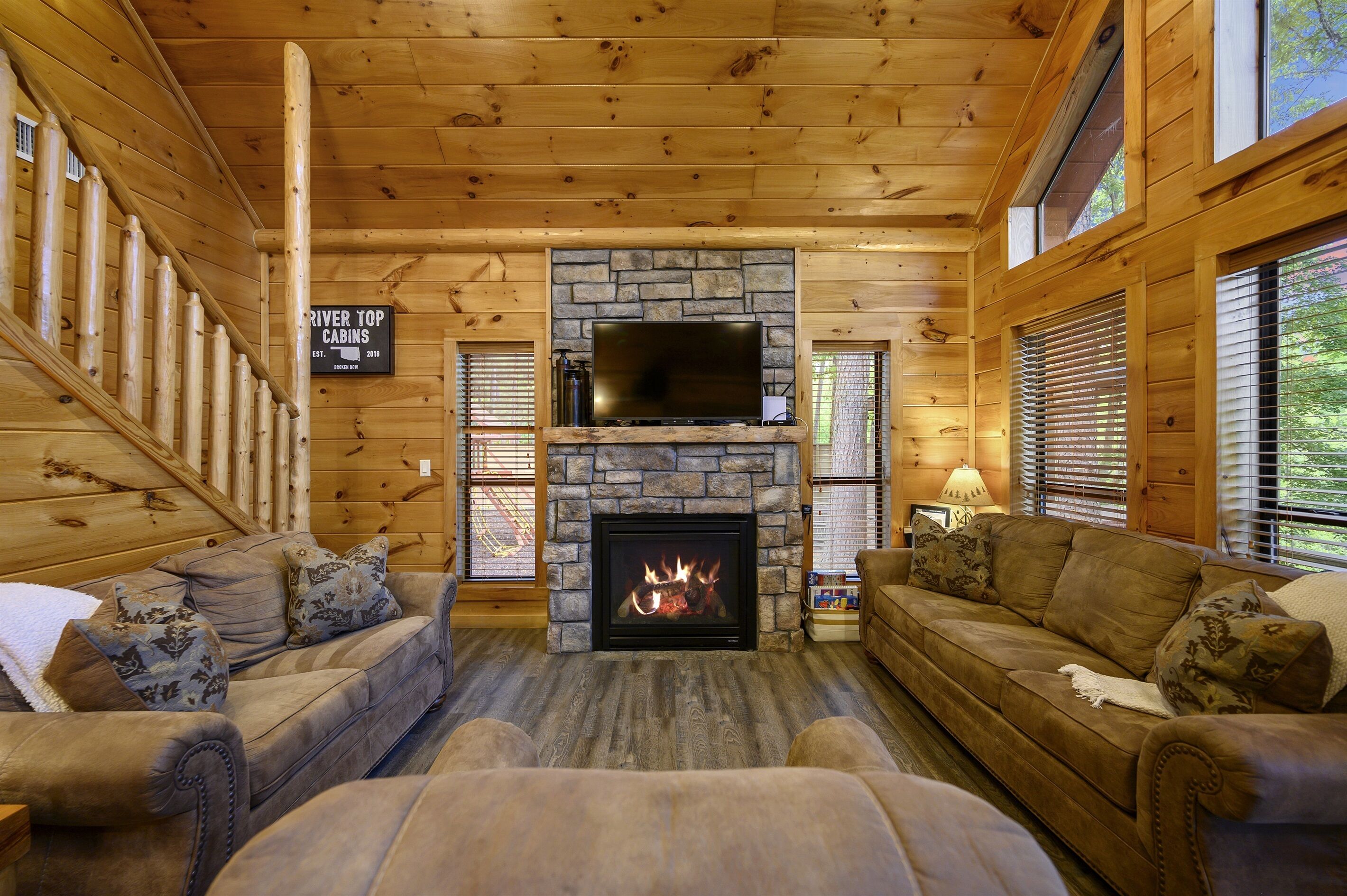 A cabin interior with a stone fireplace, wooden walls, two sofas with patterned cushions, and a staircase on the left.