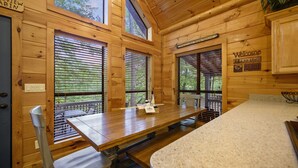 Ein Holzblockhaus-Interieur mit Esstisch, Stühlen und einer Bank. Es gibt Fenster mit Jalousien, ein Willkommensschild und ein Schild mit der Aufschrift "Cabin".