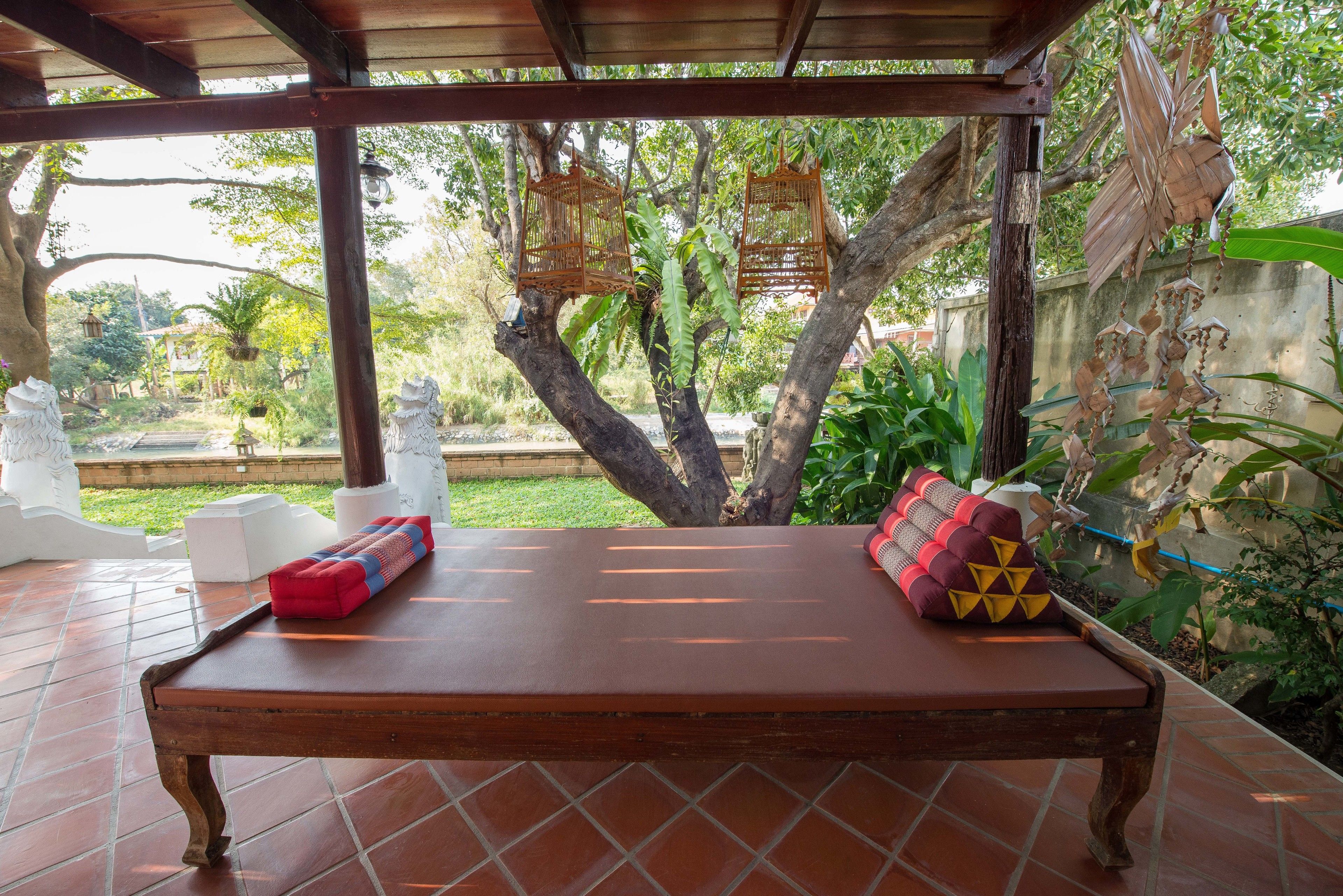 Un banco de madera con cojines en un espacio exterior cubierto, con vistas a árboles y un jardín.