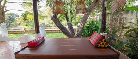 Un banco de madera con cojines en un espacio exterior cubierto, con vistas a árboles y un jardín.