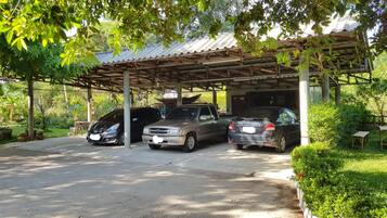 Un estacionamiento techado con tres coches aparcados bajo una cubierta metálica, rodeado de follaje y un suelo de hormigón.