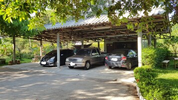 Un estacionamiento techado con tres coches aparcados bajo una cubierta metálica, rodeado de follaje y un suelo de hormigón.