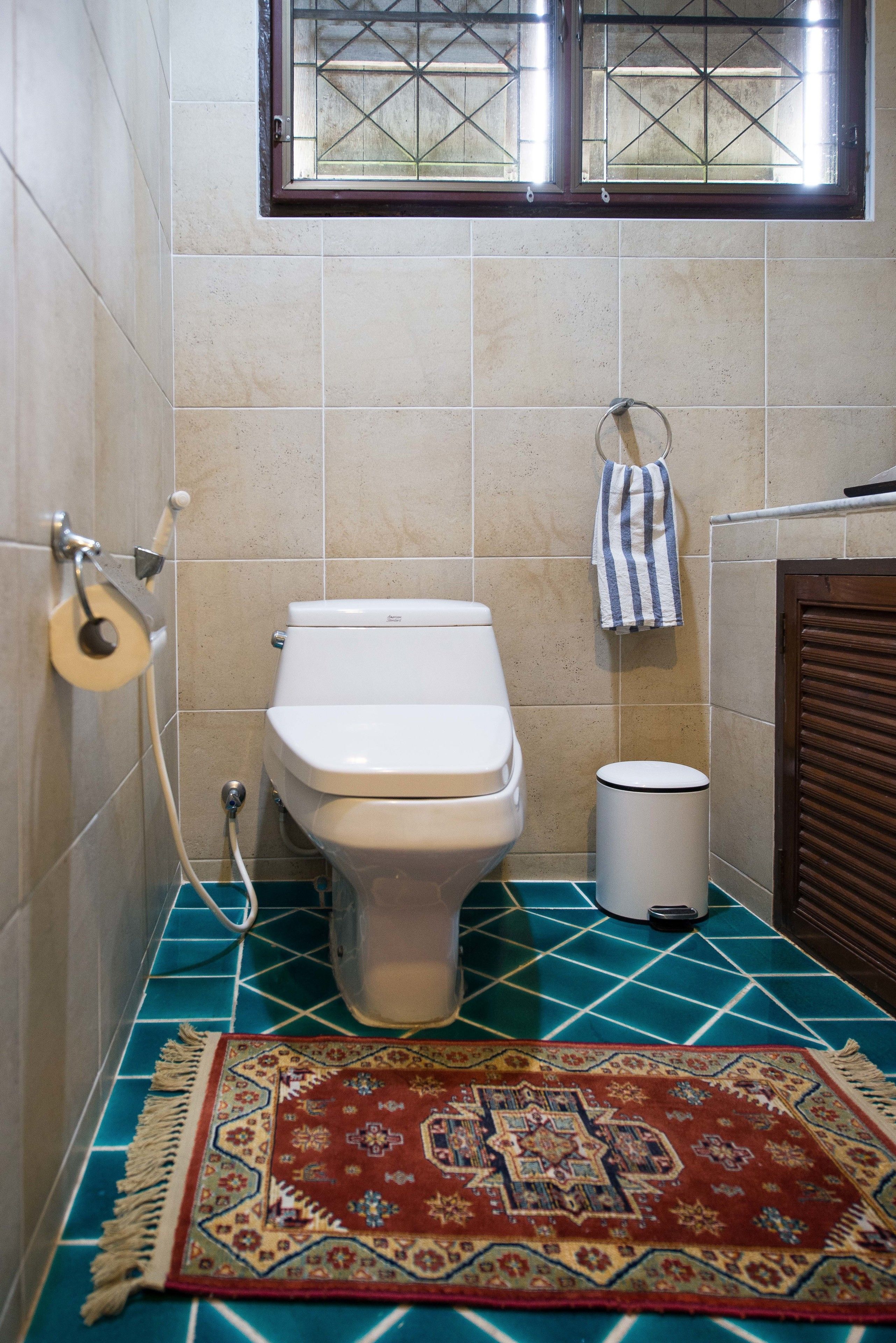 Une salle de bain avec des toilettes, une poubelle, une fenêtre et un tapis à motifs sur le sol.