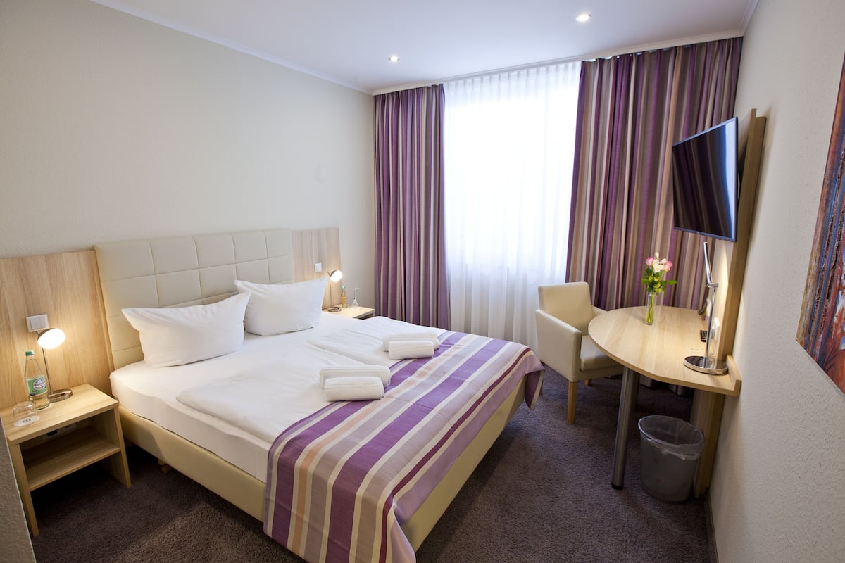 A hotel room with a bed, bedside tables, a desk, a chair, a TV, and a window with curtains.