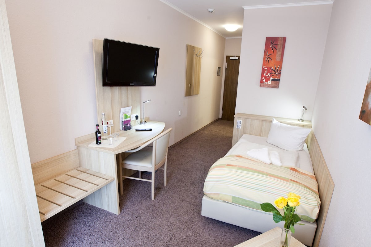A hotel room with a bed, a desk, a chair, a TV, and a small table with a vase of flowers.