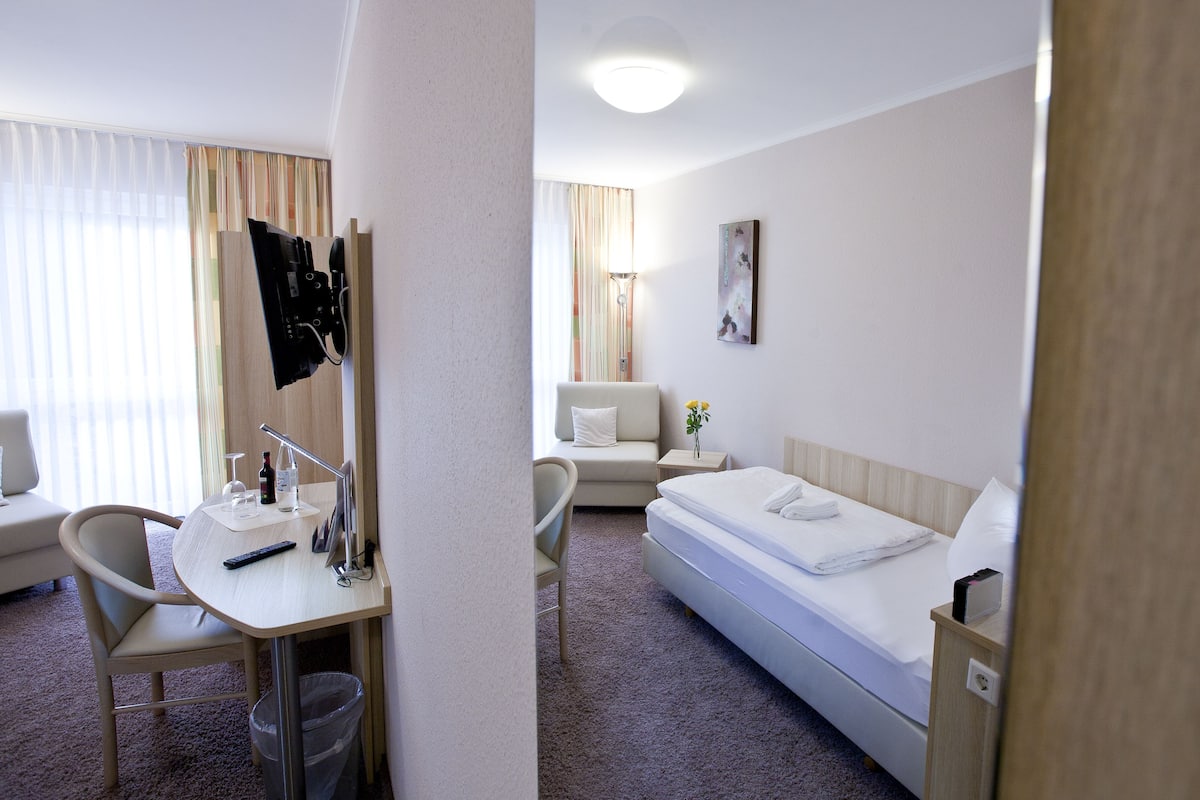 A hotel room with a bed, a desk with a chair, a small table with a lamp, a trash bin, and a window with curtains.