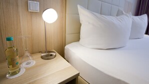 Una habitación de hotel con una cama, una mesita de noche con una botella y un vaso, y una lámpara fijada a la pared.