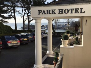 Entrada de un hotel con un cartel que dice "PARK HOTEL" y una vista de coches aparcados y una masa de agua al fondo.