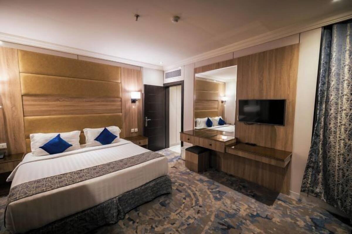 Une chambre d’hôtel avec un grand lit, une tête de lit en bois, un téléviseur à écran plat, une coiffeuse avec un miroir et une moquette à motifs.