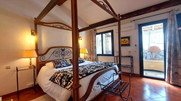 Una cama con dosel y cortinas, una mesita de noche con lámpara, una ventana con vistas al exterior y una puerta que da a un balcón.