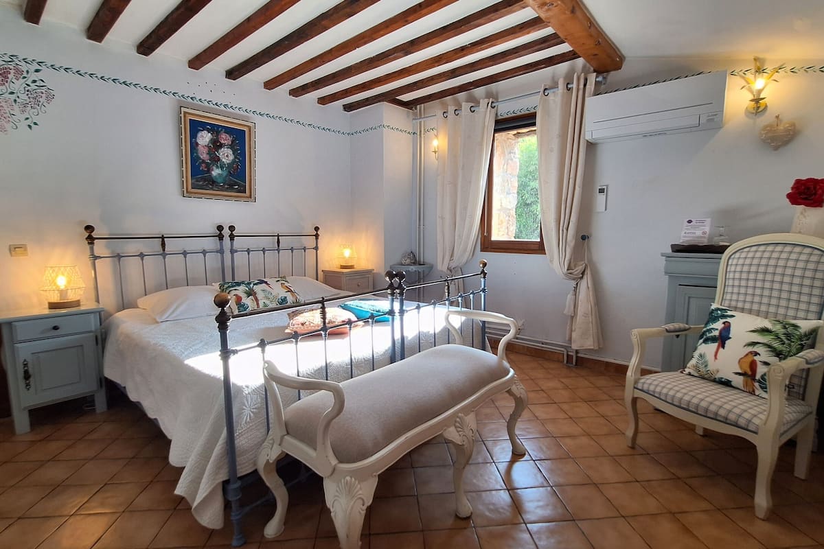 A bedroom with a bed, bedside tables, a bench, a chair, a window, and an air conditioning unit.