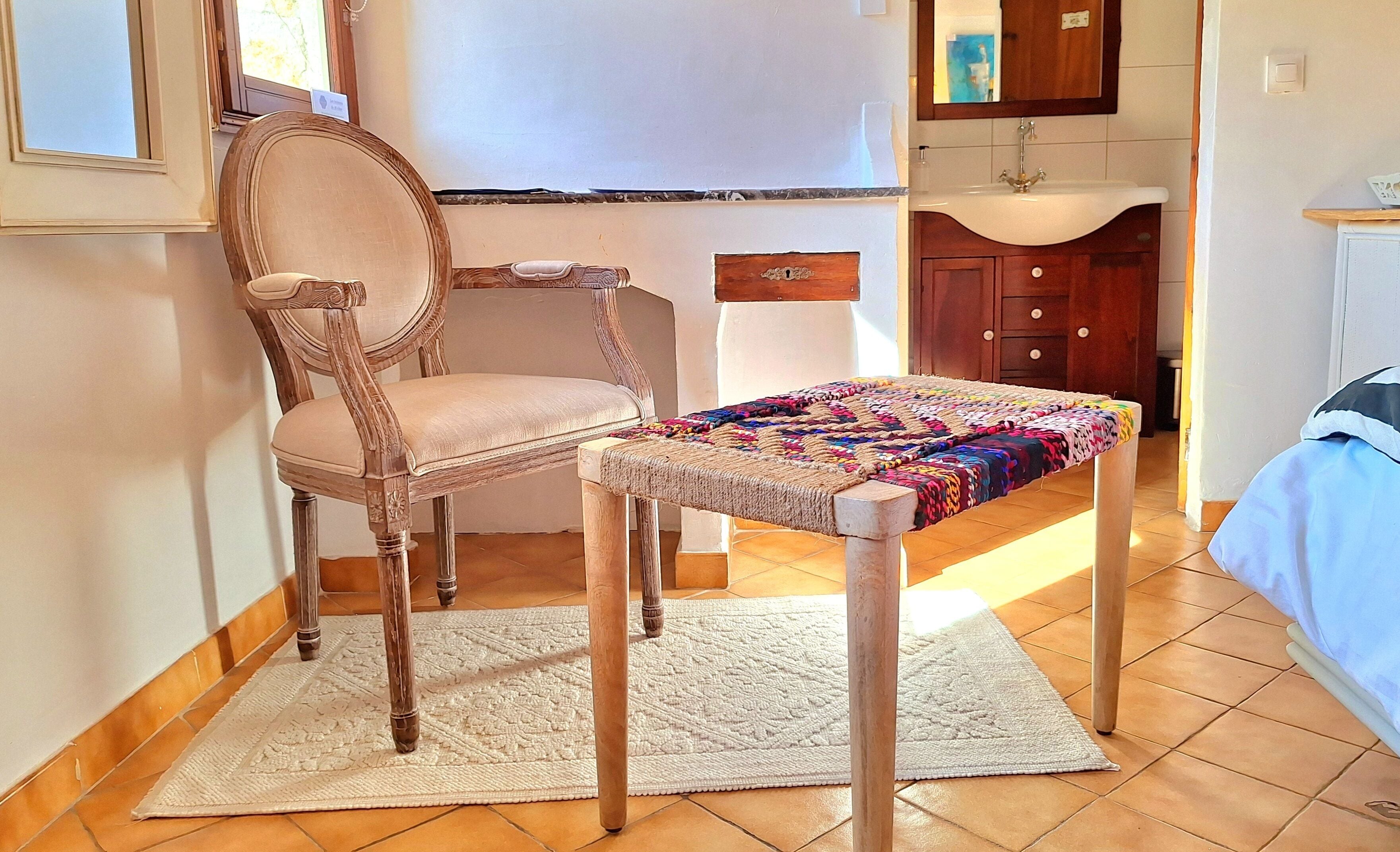 A room with a wooden chair, a small table with a colorful quilt, a white rug, a wooden cabinet, and a tiled floor.