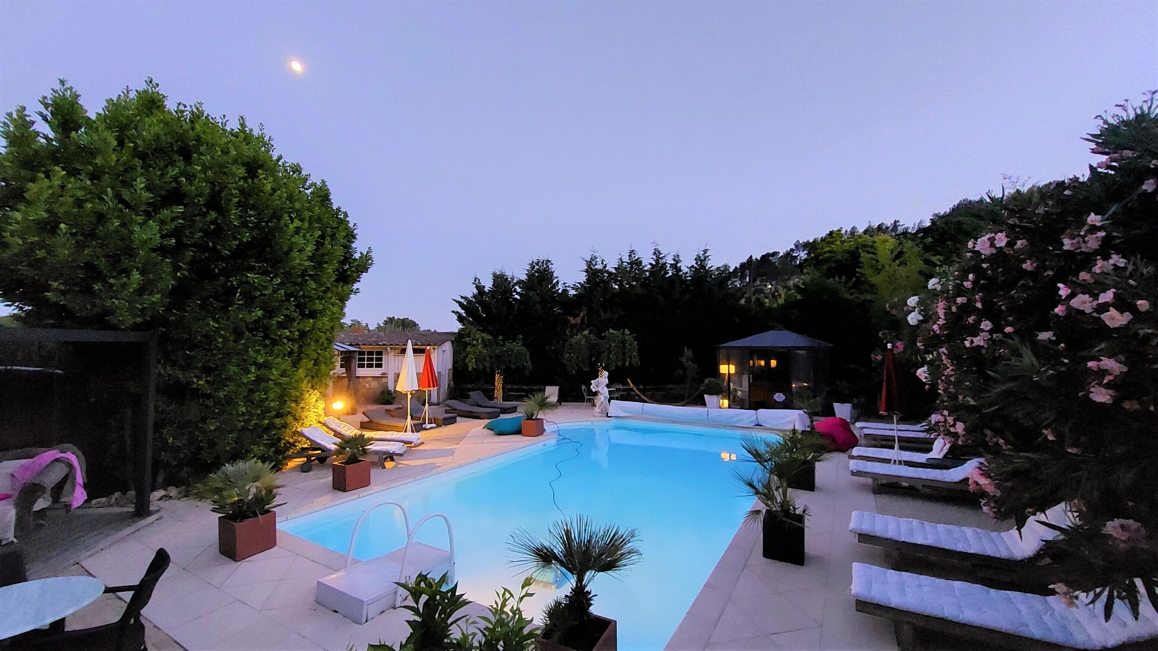 Una piscina con sillones de descanso y sombrillas, rodeada de árboles y un pabellón.
