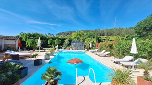 Ein Schwimmbecken mit Liegestühlen und Sonnenschirmen, umgeben von Grünpflanzen und blauem Himmel.