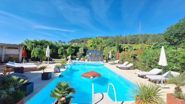 Ein Schwimmbecken mit Liegestühlen und Sonnenschirmen, umgeben von Grünpflanzen und blauem Himmel.