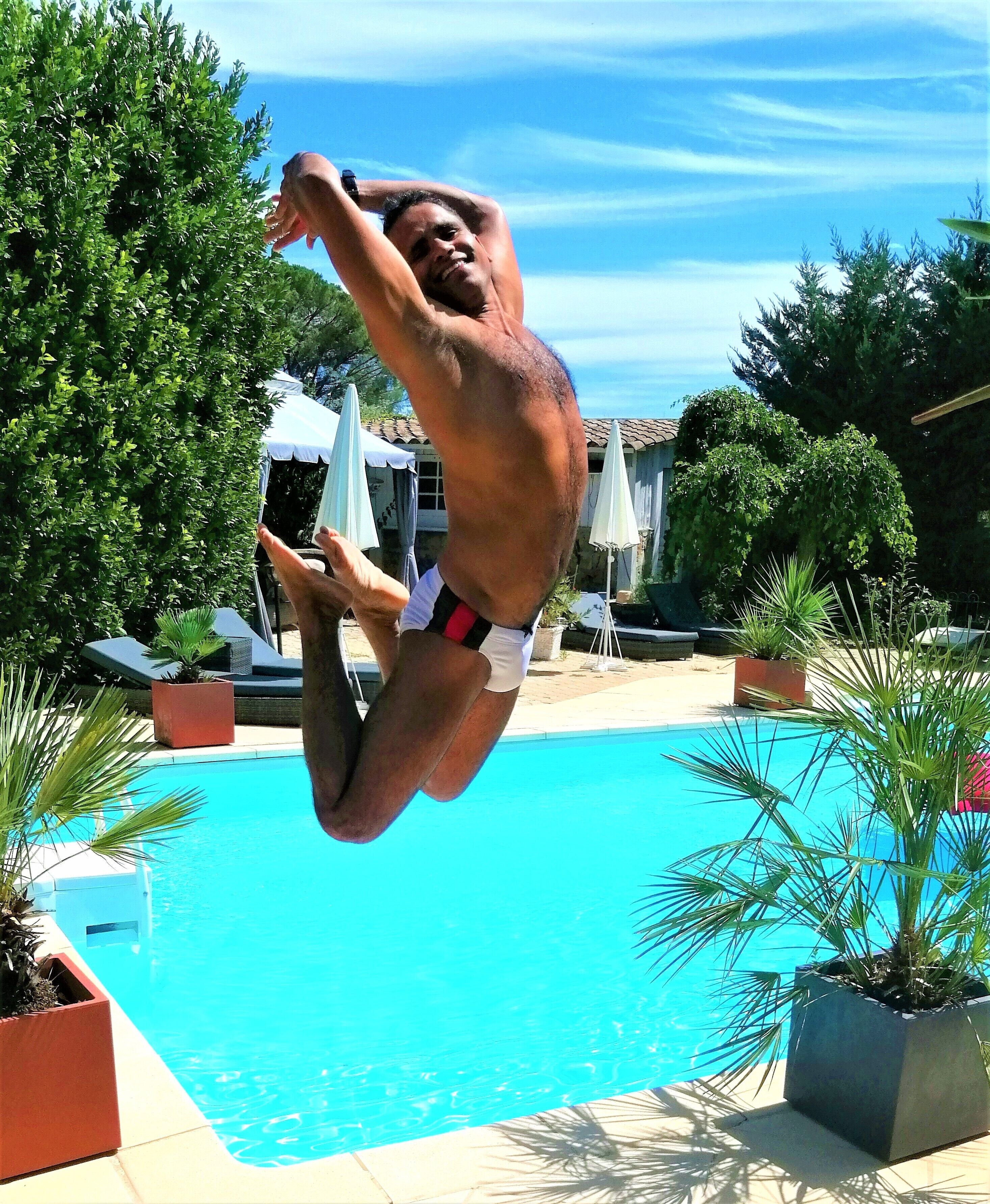 A man diving into a swimming pool surrounded by potted plants and umbrellas.