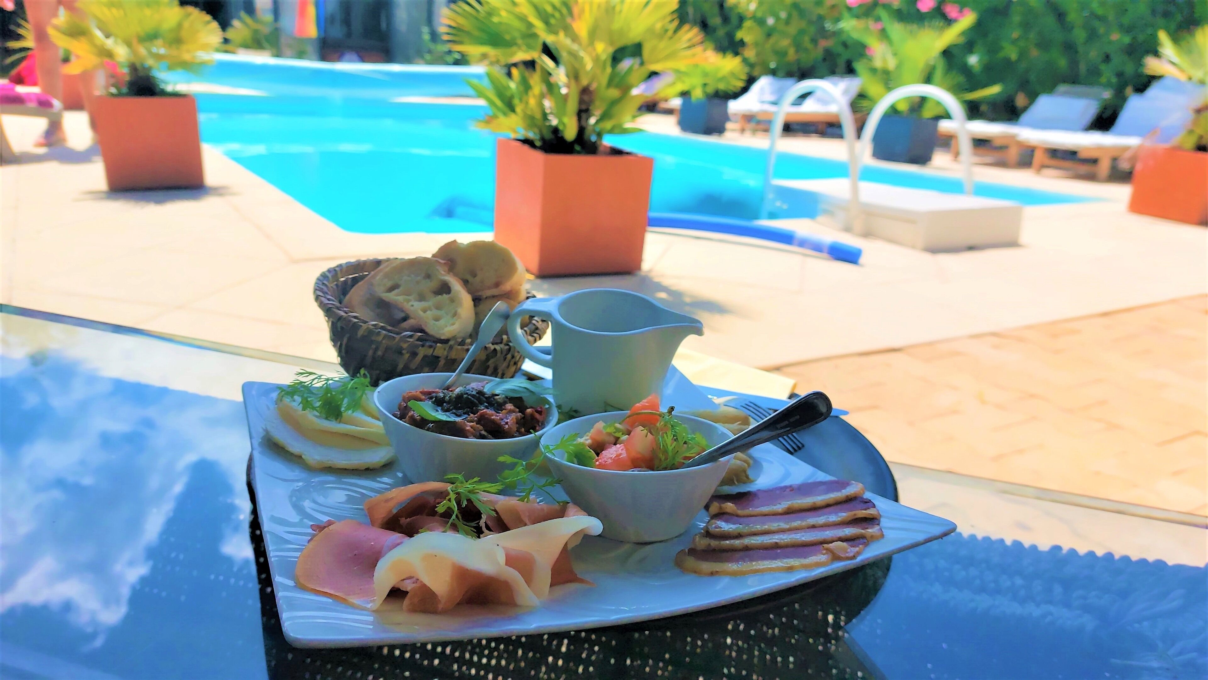 A tray with various food items and a pitcher, placed on a table near a poolside area with lounge chairs and potted plants.