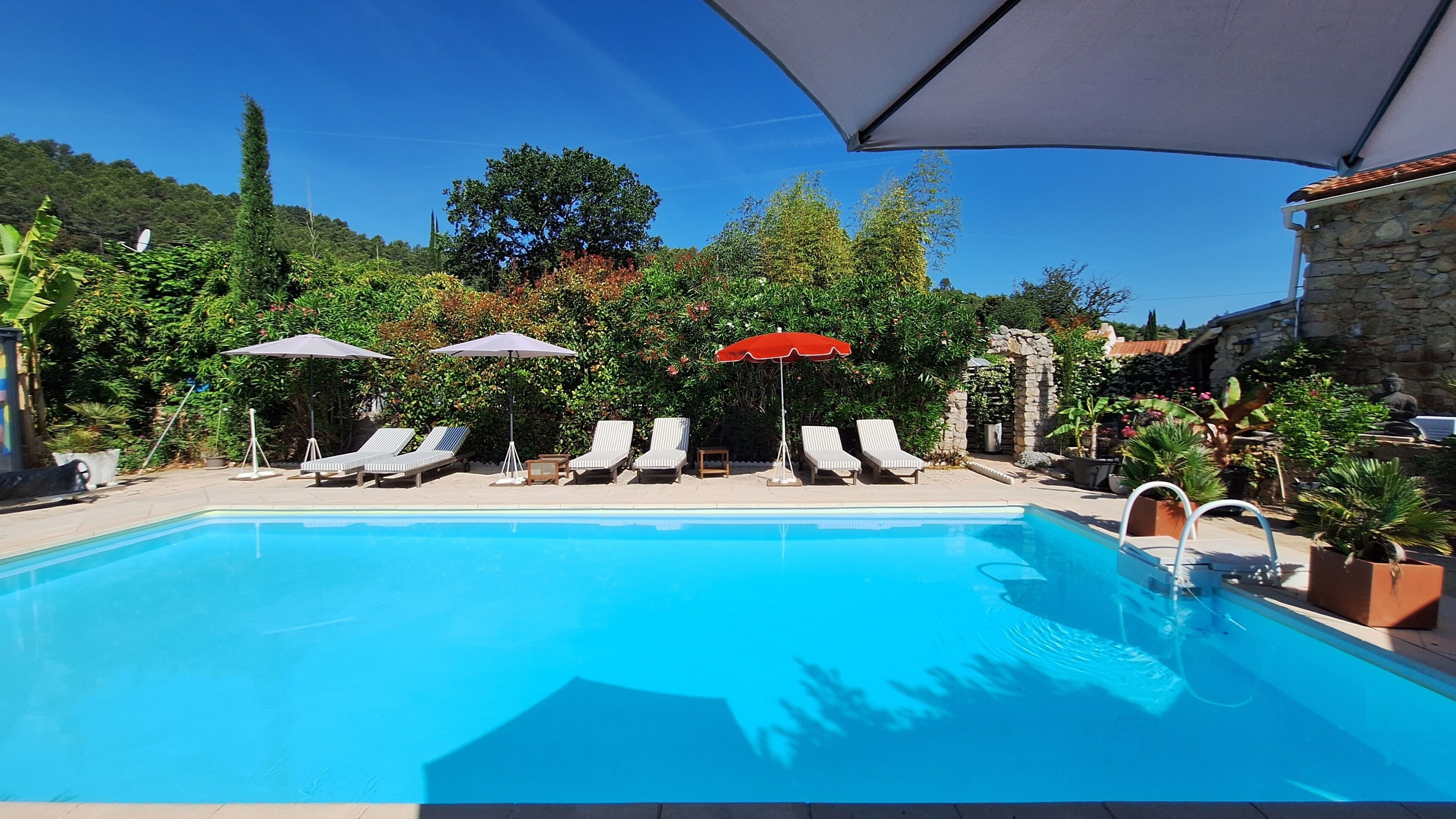 Une piscine entourée de verdure et agrémentée d’une architecture en pierre, avec des chaises longues et des parasols à proximité.