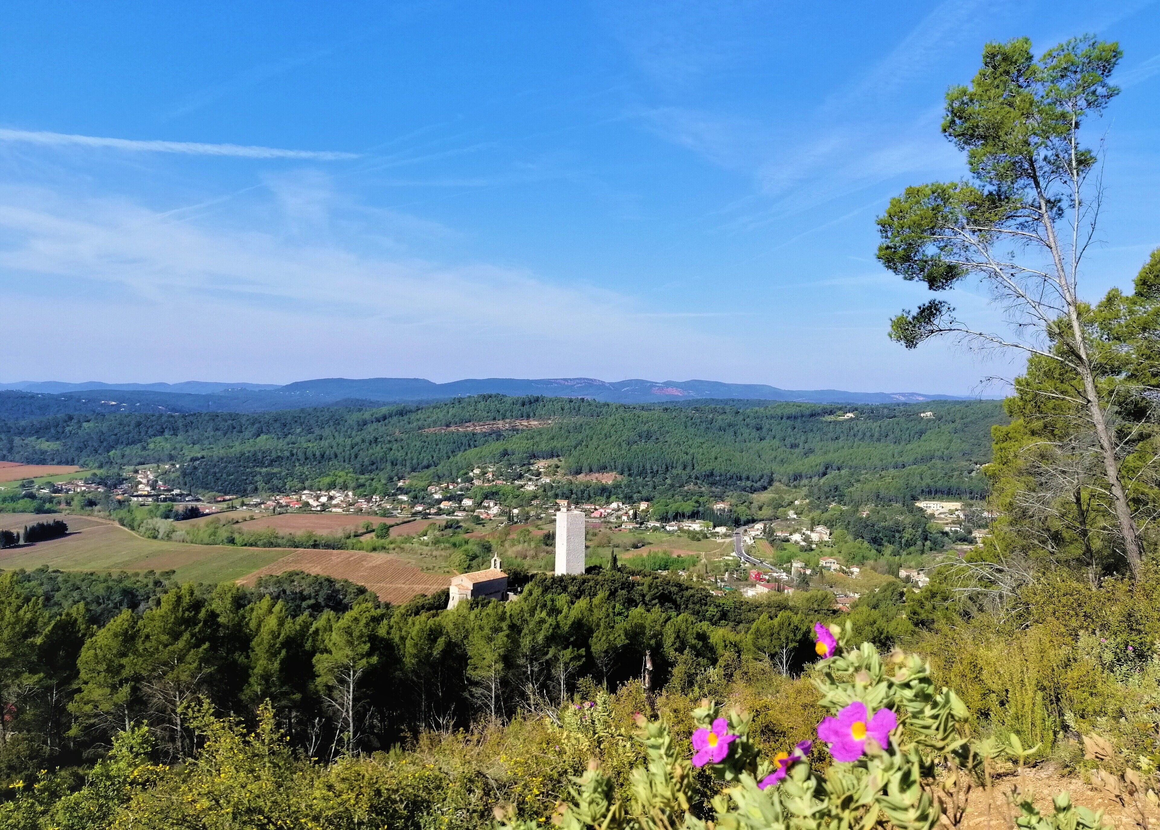 Une vue panoramique d’une ville nichée dans une vallée, avec un clocher d’église imposant, entourée d’une végétation luxuriante et de collines lointaines.