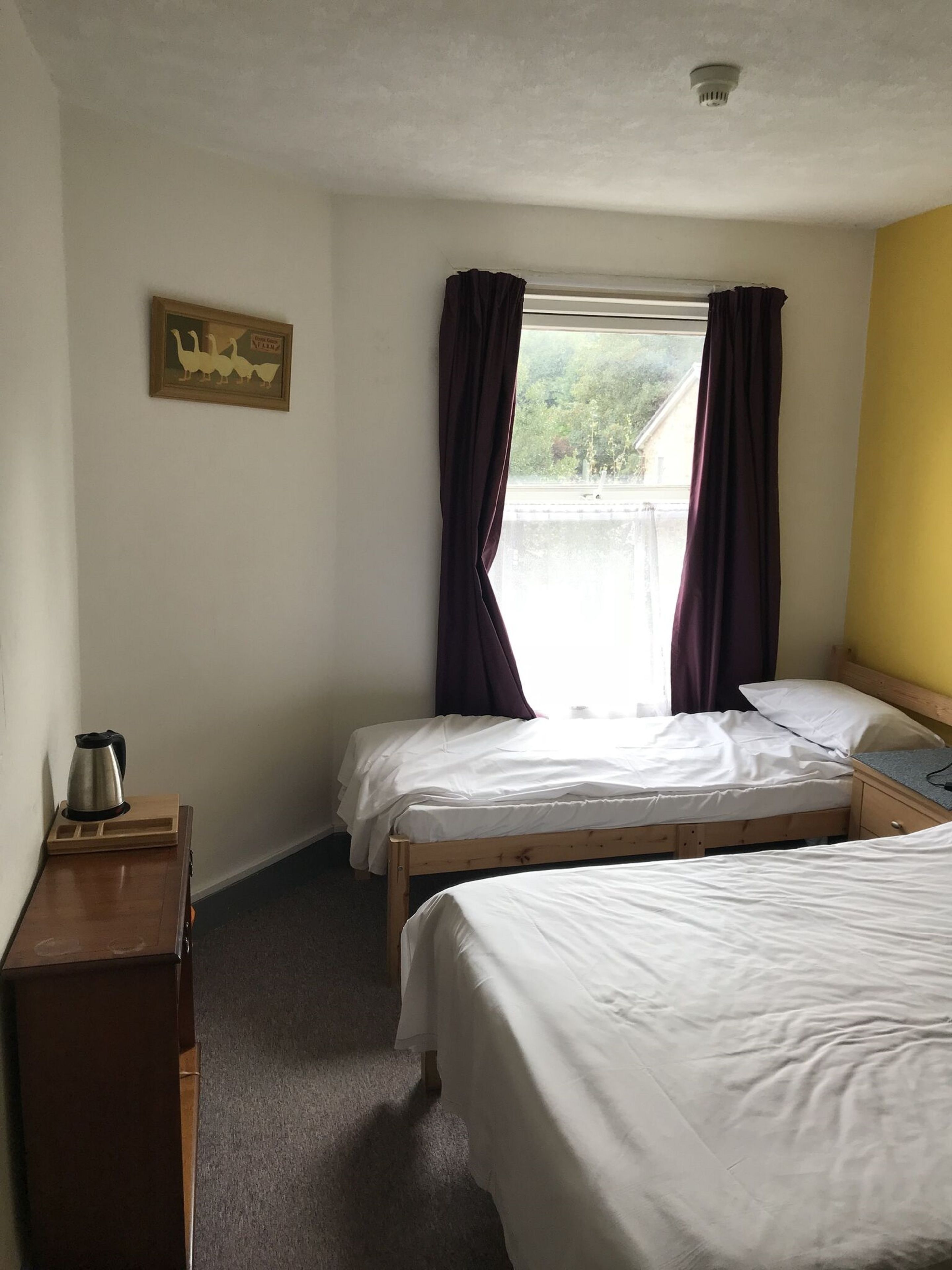 A hotel room with two single beds, a wooden nightstand, a kettle, and a window with curtains.