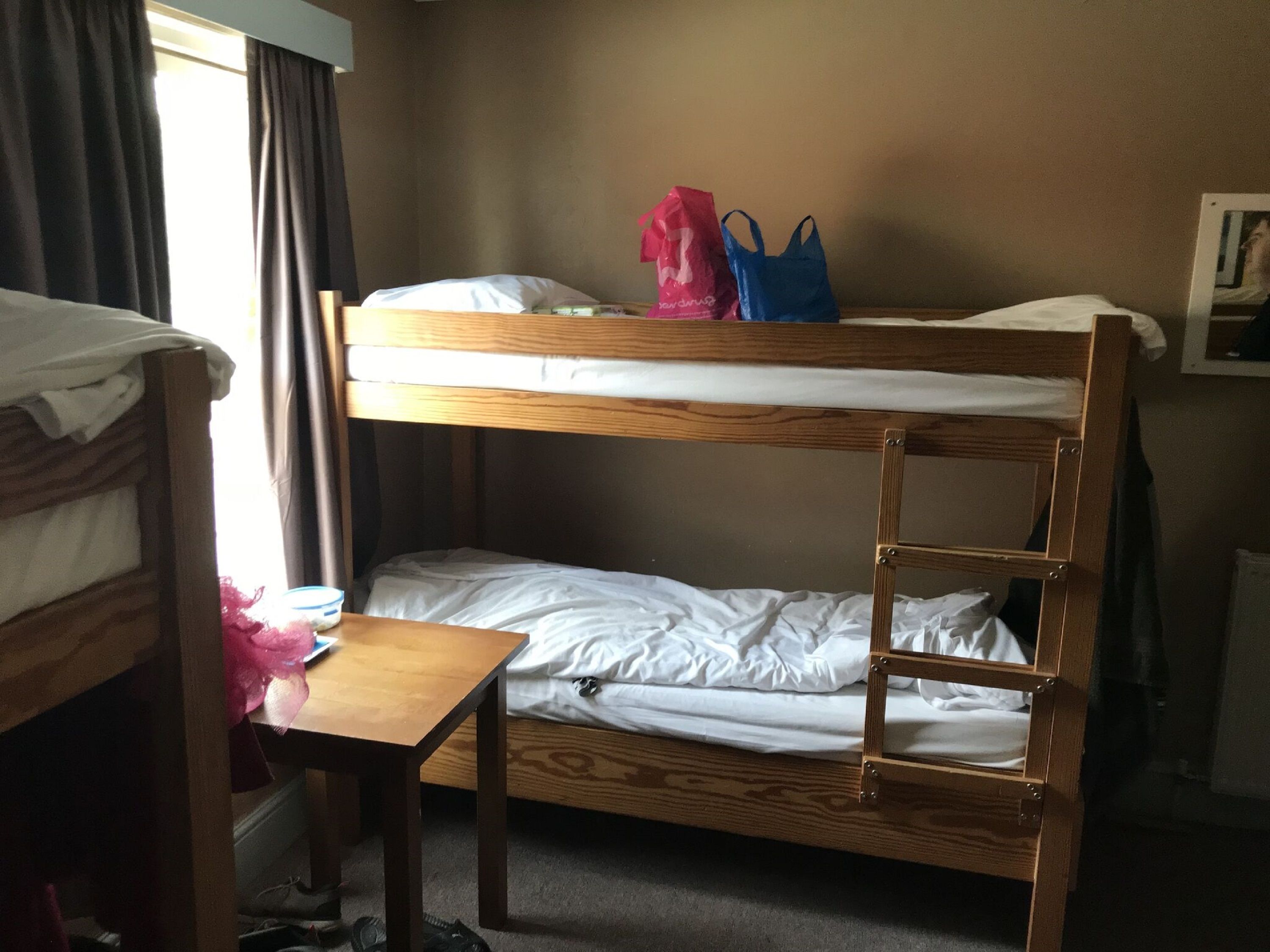 A bunk bed room with a wooden table, a pink bag, and a blue bag.