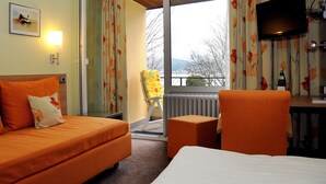A hotel room with a bed, a sofa, a chair, a desk, a television, and a window with a view of trees and a body of water. - Hotel Zur Mühle (Bad Breisig)