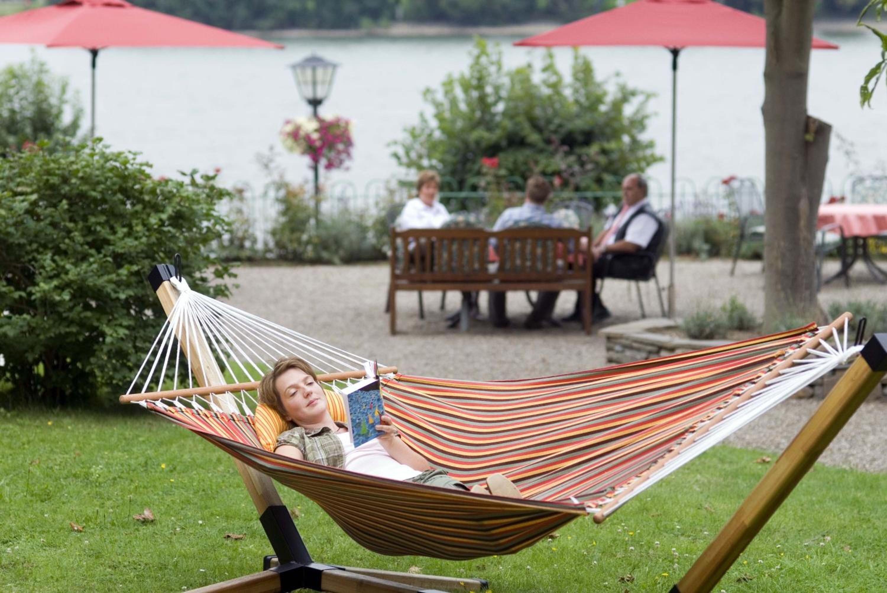 Een hangmat in een grasveld, met een meer en rode parasols op de achtergrond.