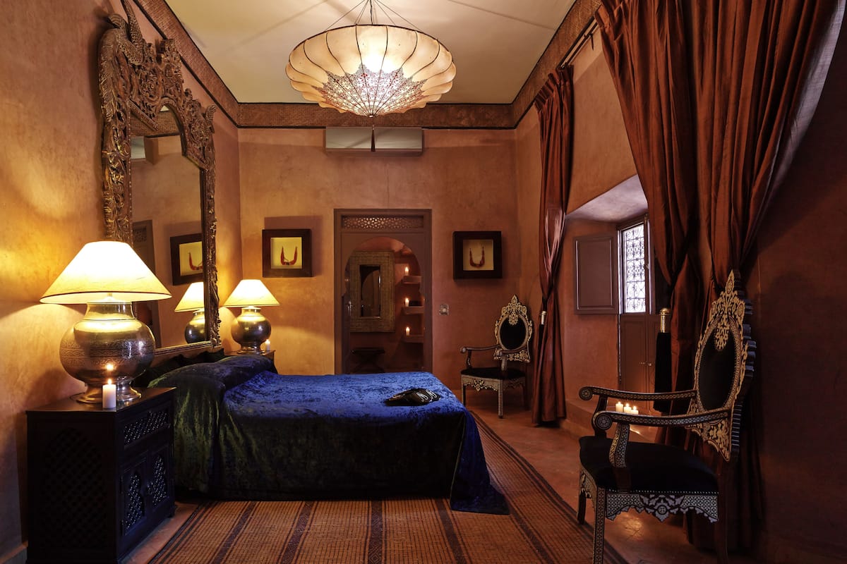 A room with a large bed, two bedside lamps, a chandelier, a mirror, and a chair.