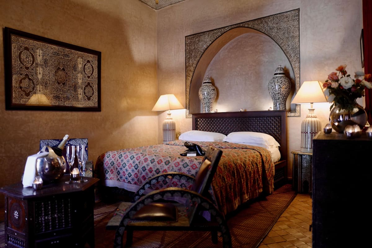 A hotel room with a large bed, two bedside lamps, a decorative mirror, a framed artwork, a small table with a vase, and a chair.