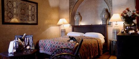 A hotel room with a large bed, two bedside lamps, a decorative mirror, a framed artwork, a small table with a vase, and a chair.
