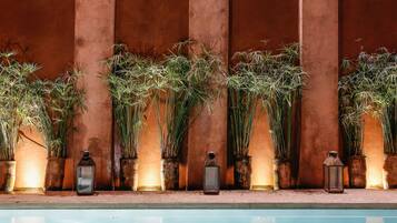 Zona junto a la piscina con plantas altas y faroles frente a un muro con textura.