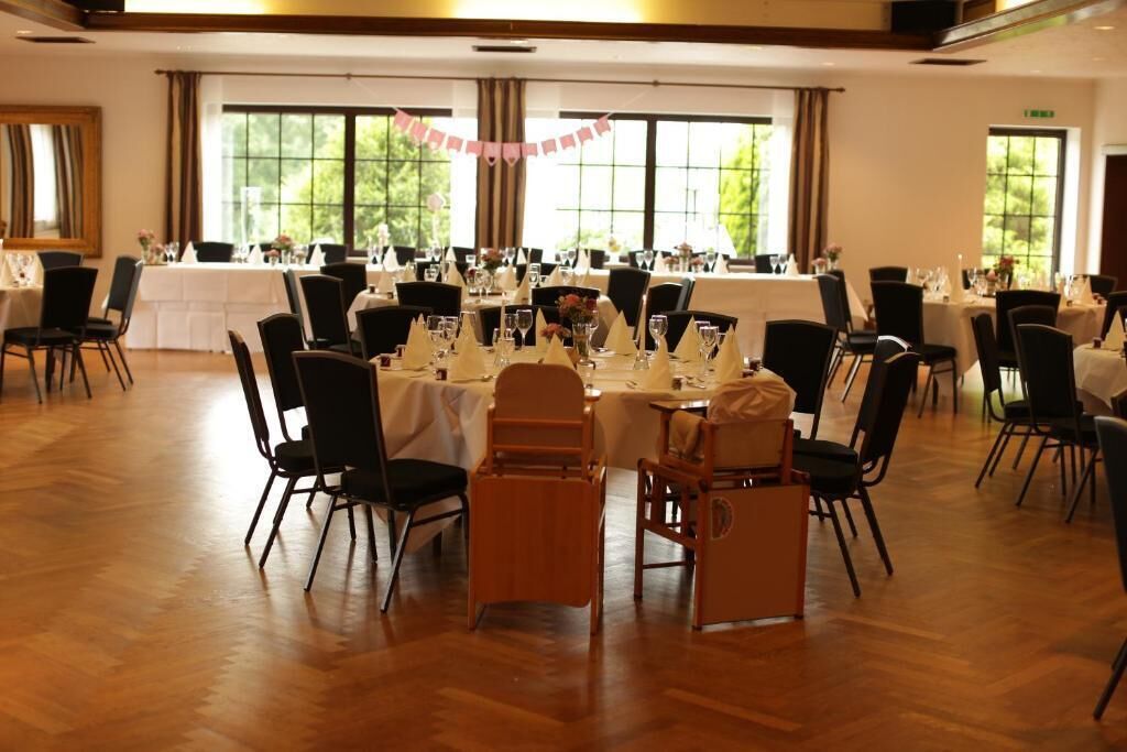 Une salle de banquet avec des tables rondes dressées pour le repas, des chaises noires et de grandes fenêtres laissant entrer la lumière naturelle.