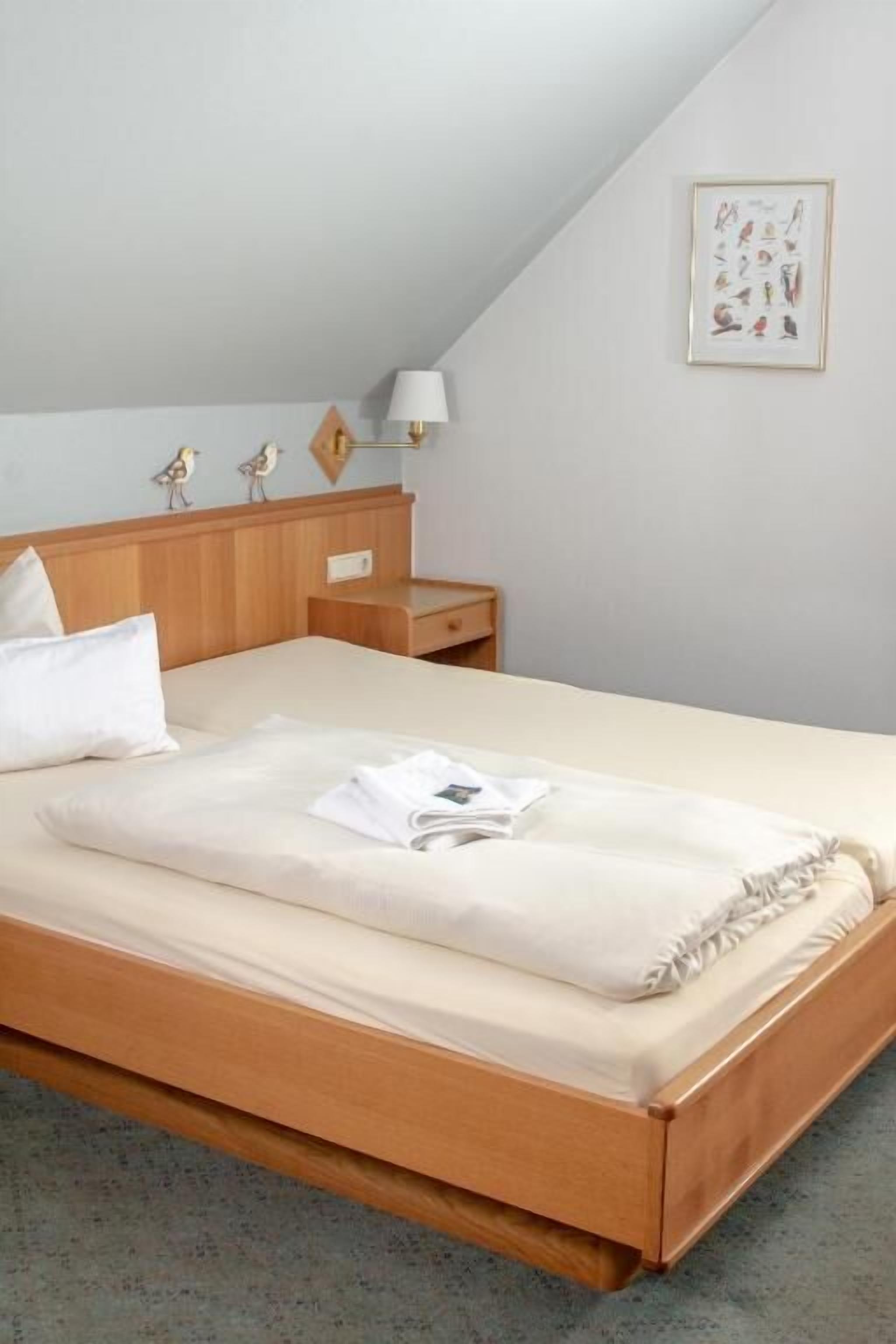 A neatly made bed with a white comforter and pillows, a wooden headboard, a bedside table with a lamp, and a framed picture on the wall.