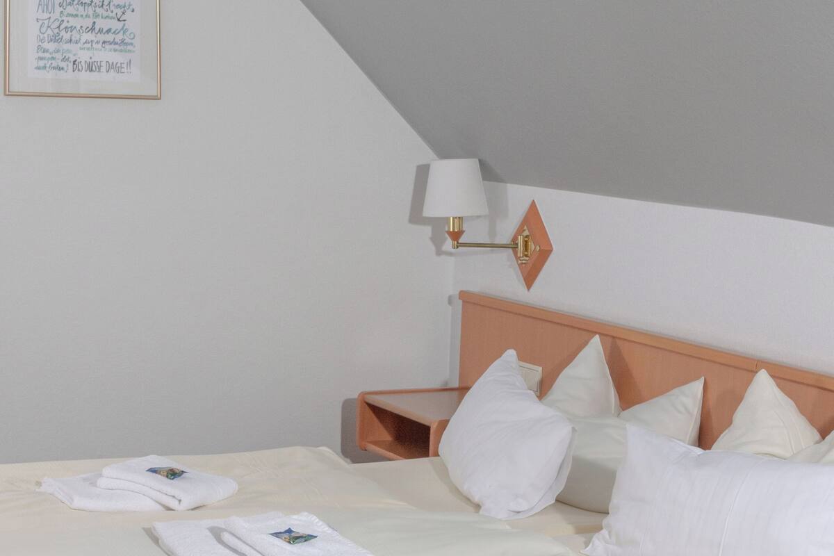 En pænt redt seng med hvide sengetæpper og puder, et natbord med en lampe, og en indrammet seddel på væggen.