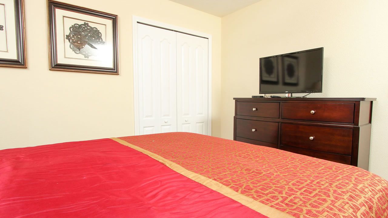 Un dormitorio con colcha roja, cómoda de madera, televisor y puerta blanca.