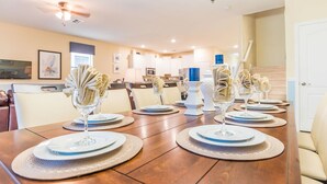 A dining table set with plates, glasses, and napkins, surrounded by chairs in a well-lit room with a kitchen in the background. - Grhrfd1469 - Champions Gate Resort - 9 Bed 5 Baths House (Davenport)
